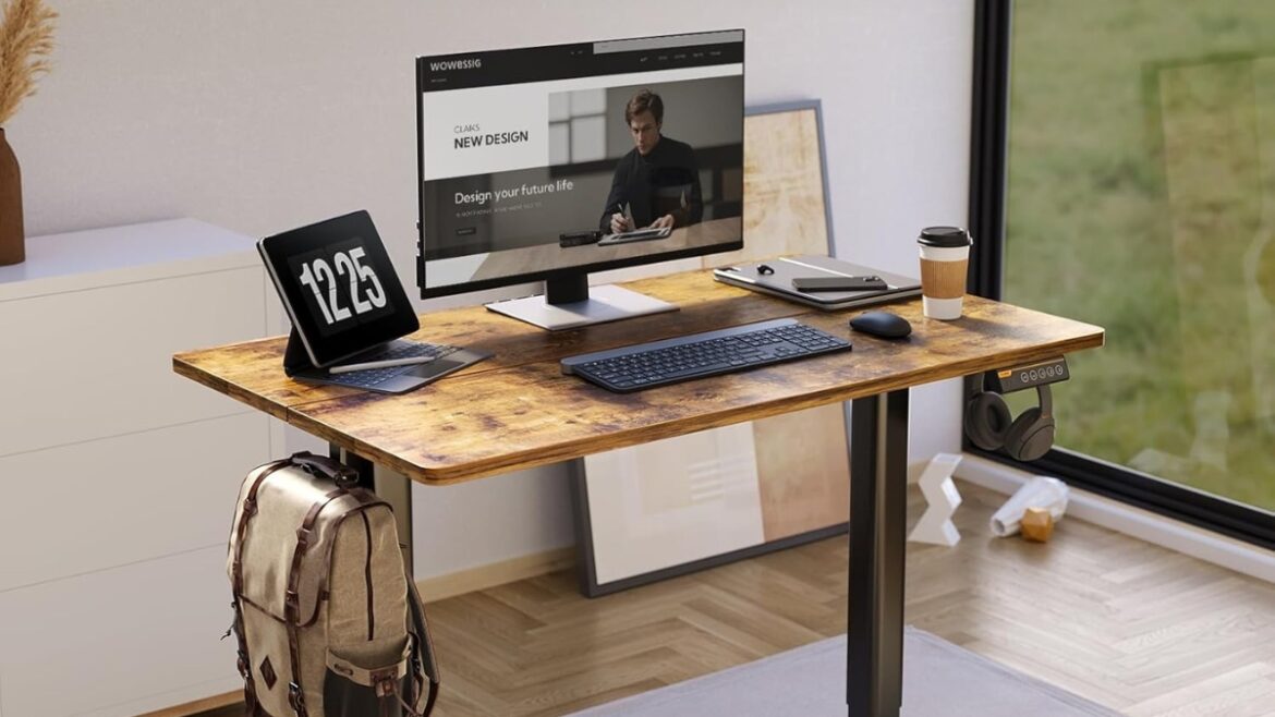 A Claiks Electric Standing Desk with a rustic wood top holding a monitor, keyboard, tablet, and coffee cup in a bright home office.