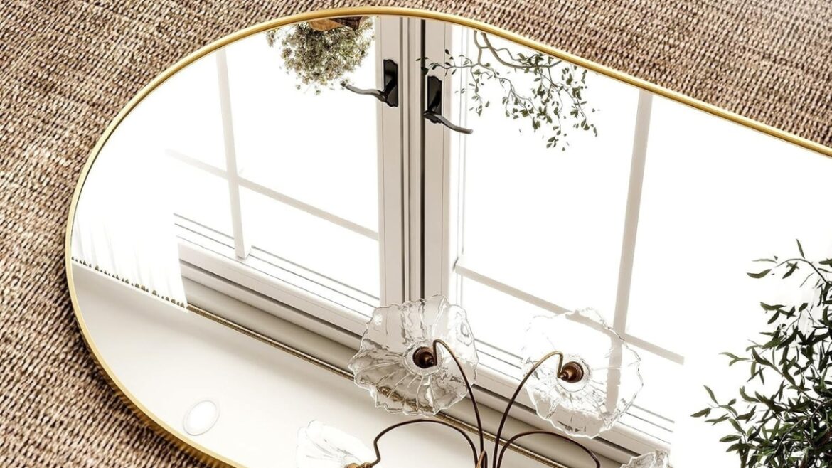 Gold arched mirror on a textured surface reflecting a window, light fixture, and green plants.