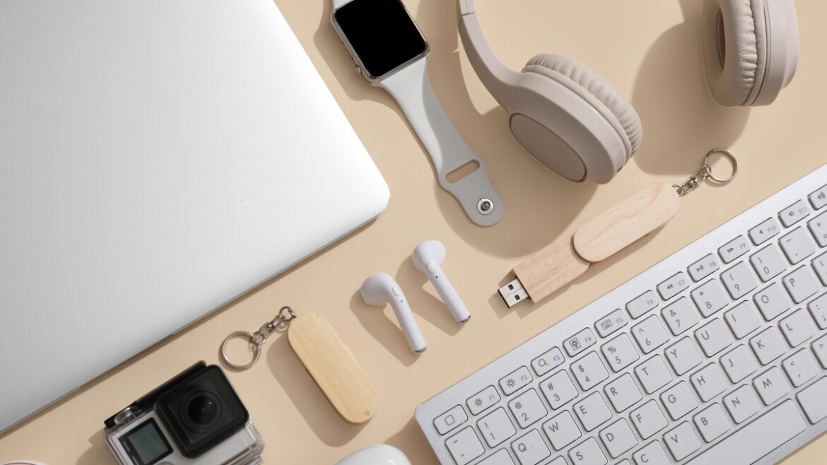 Flat lay of modern electronic gadgets including laptop, wireless headphones, smartwatch, keyboard, earbuds, USB drives, and action camera on beige background.