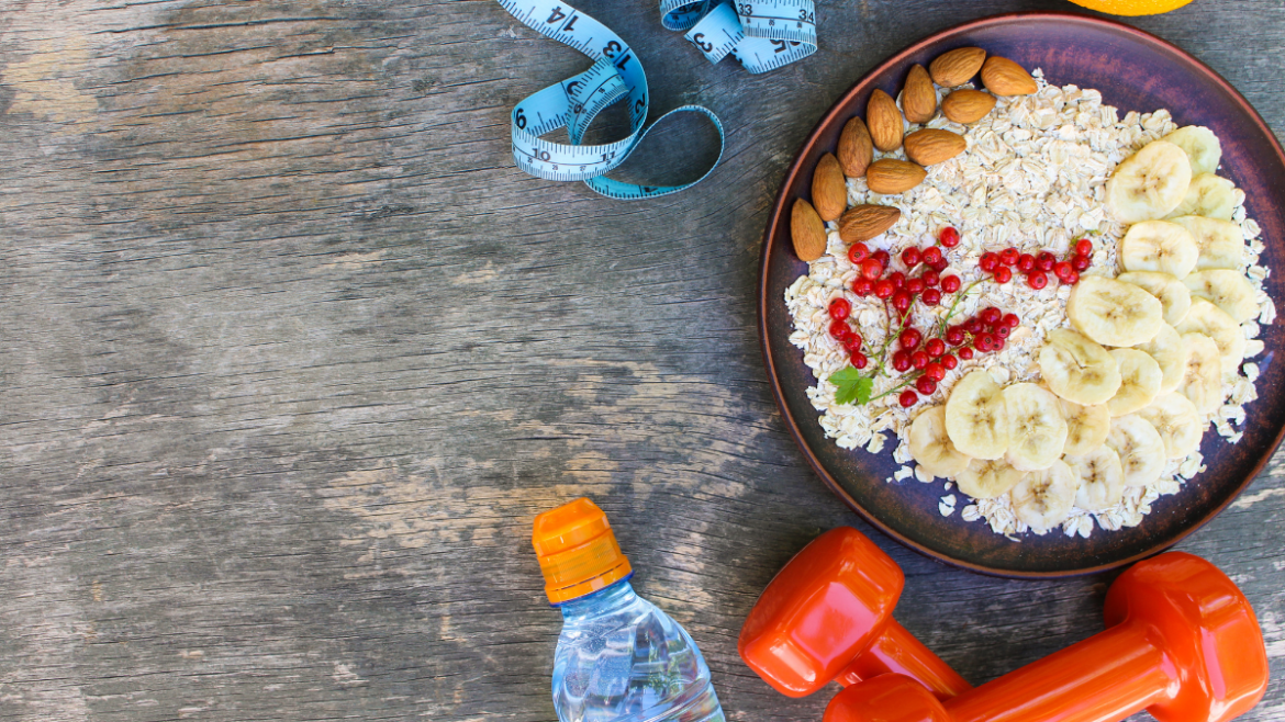 Sports nutrition setup with oatmeal, bananas, almonds, berries, measuring tape, water bottle, and dumbbells on a wooden table.